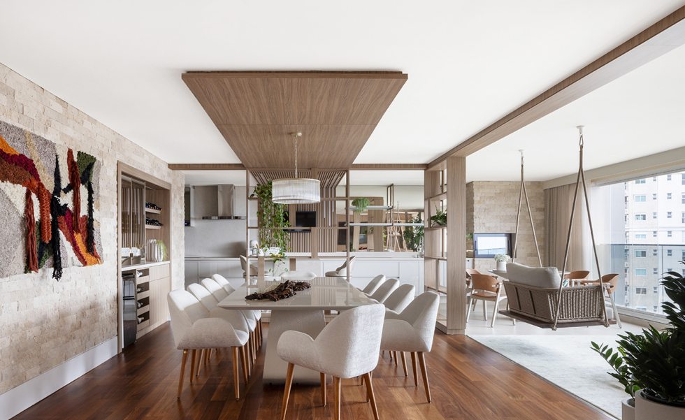 No jantar, a tapeçaria assinada por Marilene Ropelato dialoga com o painel de madeira sobre a mesa da Masotti e as cadeiras da Sierra. O conjunto destaca o uso de texturas naturais e reforça a conexão com a cozinha e a varanda com churrasqueira.