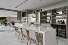 A sala de jantar, integrada com a churrasqueira gourmet, possui poltronas em linho e madeira para acomodar doze pessoas sentadas. A churrasqueira traz bancada em silestone branco com mais seis lugares, para acomodar um número maior de convidados com conforto